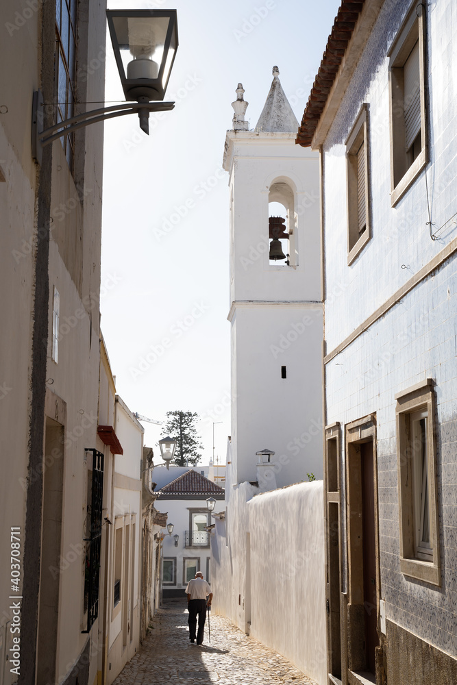 Elderly man walking with a cane in a street with white houses. High part of the town of Tavira, Portugal. Small towns of the Algarve.