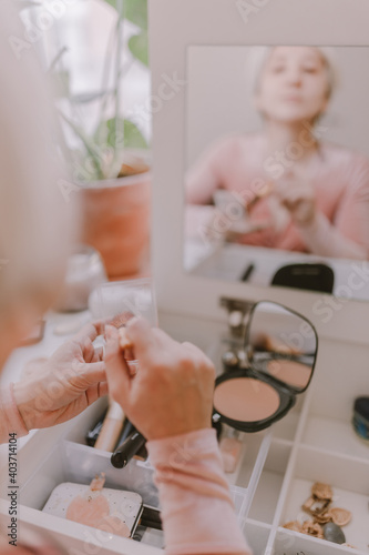 Young millennial woman is dressing up. Wearing earrings. Beauty routine. Make up artist. Mascara, lipstick, blushes. Pastel colors. Dressing vanity mirror. Pretty blonde lady. White bedroom light.