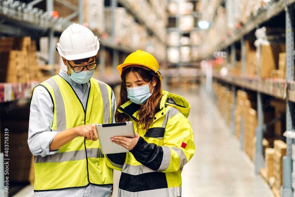 Asian engineer man and woman in helmets in quarantine for coronavirus wearing protective mask working in new normal at shelves with goods background in warehouse.logistic and business export Asian engineer man and woman in helmets in quarantine for coronavirus wearing protective mask working in new normal at shelves with goods background in warehouse.logistic and business export