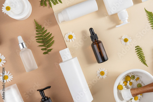 Set of care cosmetics. Various bottles, tubes with cosmetic, chamomile flowers, fern leaves and mortar bowl with pestle on beige and brown background. Beauty concept. Top view Flat lay