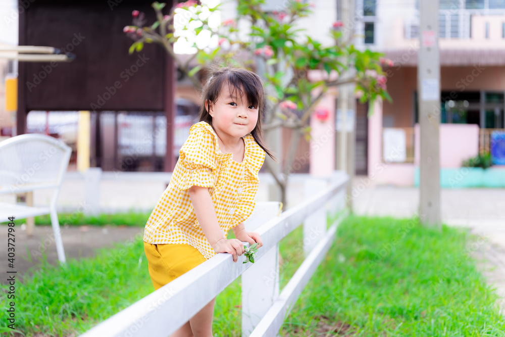 Fototapeta premium Little girl in a yellow dress stood by the white fence. Child pretended to be climbing the fence to the other side. Children smiled sweetly and looked at the camera. Kid are 5 years old.