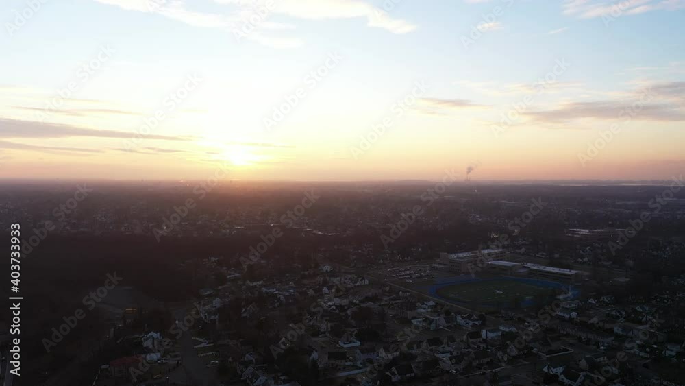 An aerial shot over a suburban neighborhood at sunrise. The drone camera truck right and pan left while looking at the colorful and cloudy sky. The neighborhood below is dark. An aerial shot over a suburban neighborhood at sunrise. The drone camera truck right and pan left while looking at the colorful and cloudy sky. The neighborhood below is dark.
