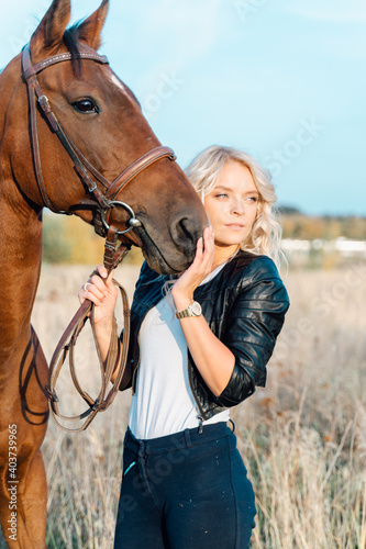 A pretty blonde in a black jacket stands in a field next to a horse at sunset.
