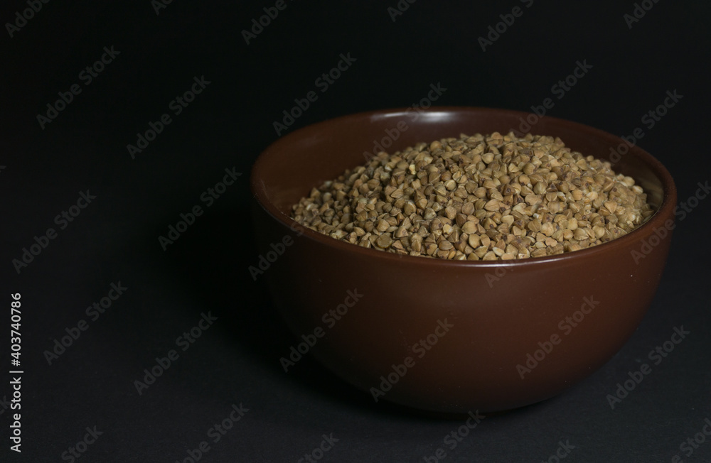 Brown uncooked buckwheat in a bowl with scattered grains nearby on a black background Brown uncooked buckwheat in a bowl with scattered grains nearby on a black background