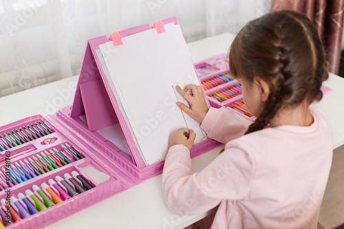 Fényképezés Little assiduous female kid sitting at table, using ruler for drawing tree, painting with set of art and creativity, posing backwards to camera, having pigtails