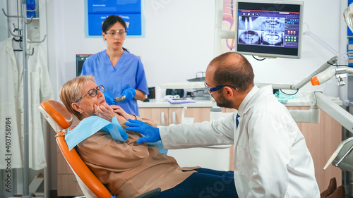 Retired woman having symptoms of gums pain holding hand on cheek while talking with doctor. Elderly patient explaining dental problem to doctor indicating mouth while nurse preparing sterile tools.