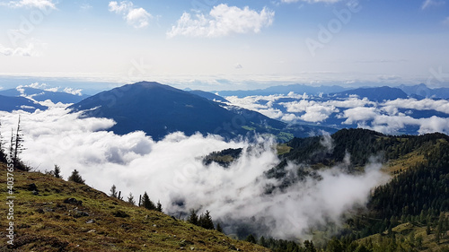 A panoramic view on the Alpine valley from the top of Granattor in Austrian Alps. The valley is shrouded in fog. The slopes are lush green. Many mountain poking above the clouds. Mystery mood