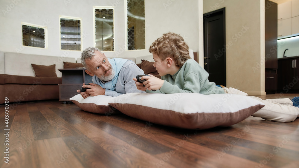 Grandfather with grandson lying on pillows on the floor and playing video games, spending time together at home
