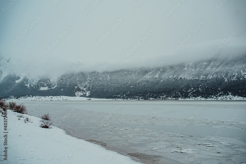 Frozen lake in winter mountains. It is snowing. The hills of pines. Frozen lake in winter mountains. It is snowing. The hills of pines.