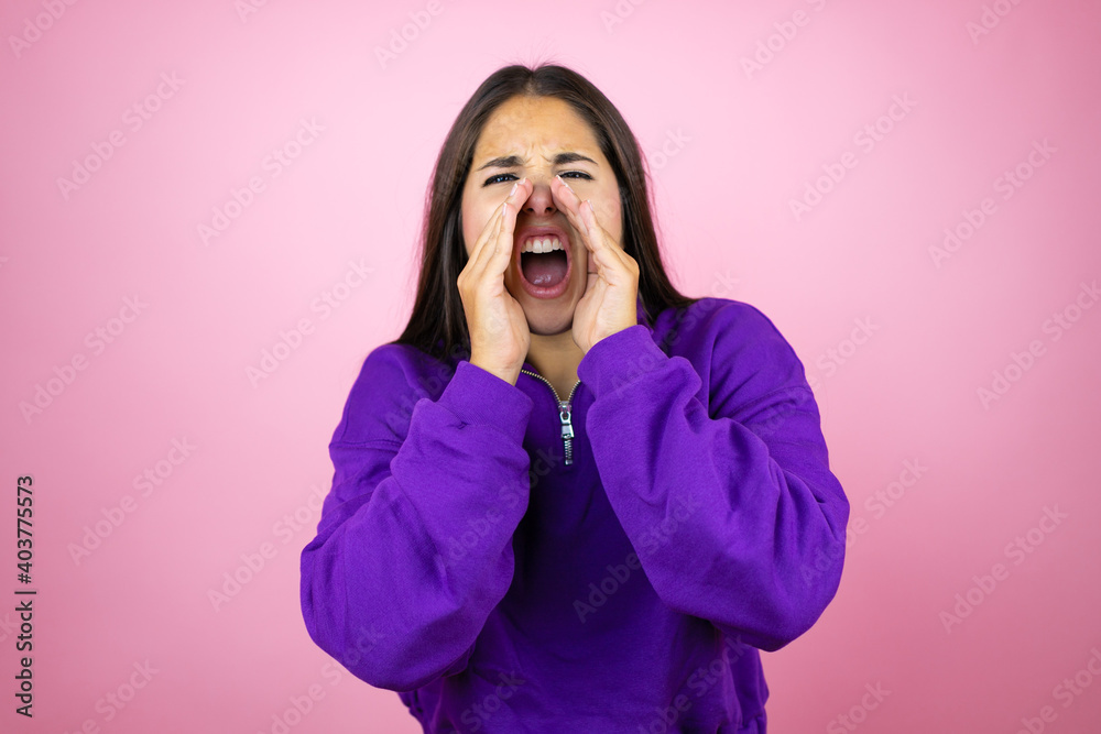 Young beautiful woman wearing sweatshirt over isolated pink background shouting and screaming loud to side with hands on mouth Young beautiful woman wearing sweatshirt over isolated pink background shouting and screaming loud to side with hands on mouth