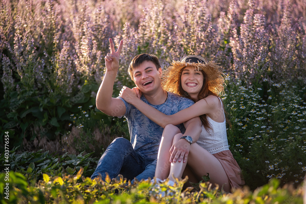 Fototapeta premium Happy couple in love sits hugging by blooming sage field. A young man with beautiful girl in straw hat are having fun, fooling around, waving their hands, showing their tongue. travel of crazy lovers