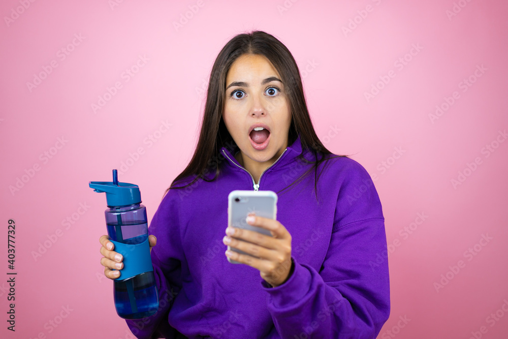 Young beautiful woman wearing sweatshirt holding a water bottle over isolated pink background chatting on the phone with a surprised expression Young beautiful woman wearing sweatshirt holding a water bottle over isolated pink background chatting on the phone with a surprised expression
