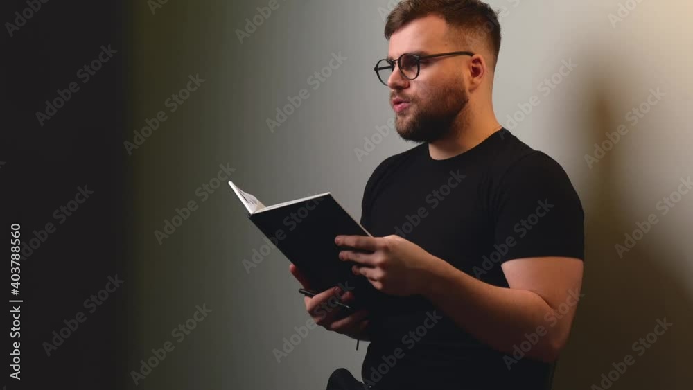 Bearded boy is reading something from the book to somebody sitting on a gray studio wall wearing glasses and black t-shirt Bearded boy is reading something from the book to somebody sitting on a gray studio wall wearing glasses and black t-shirt