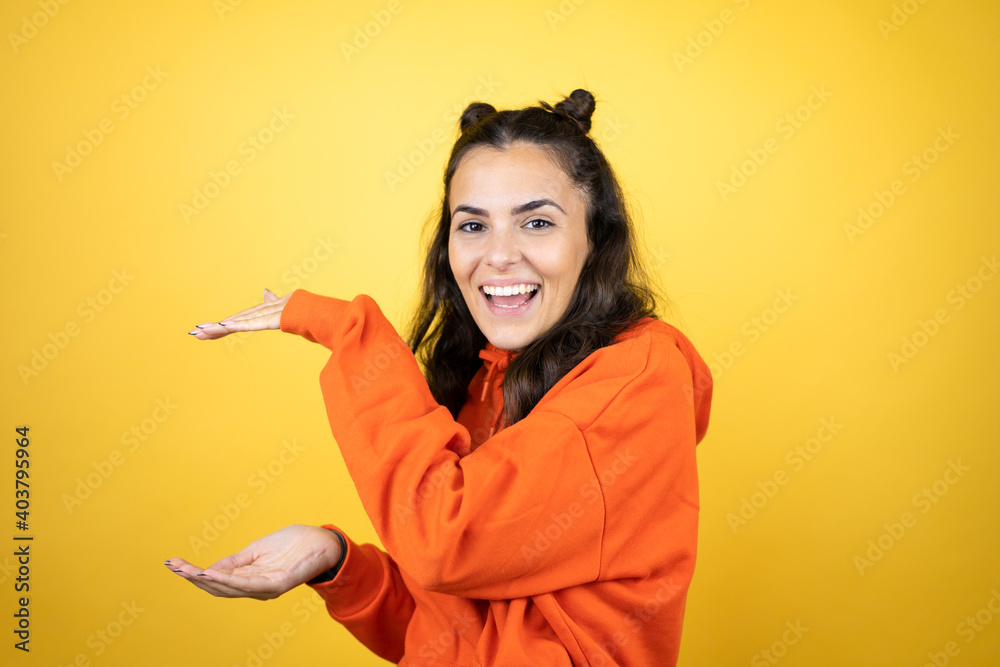Young beautiful woman wearing sweatshirt over isolated yellow background gesturing with hands showing big and large size sign, measure symbol. Smiling . Measuring concept.
