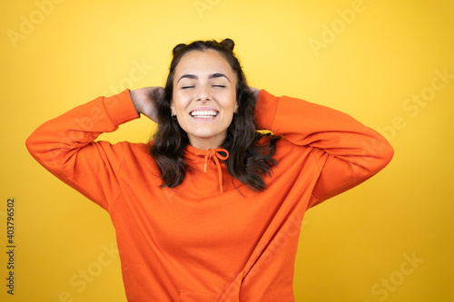 Young beautiful woman wearing sweatshirt over isolated yellow background relaxing and stretching, arms and hands behind head and neck smiling happy