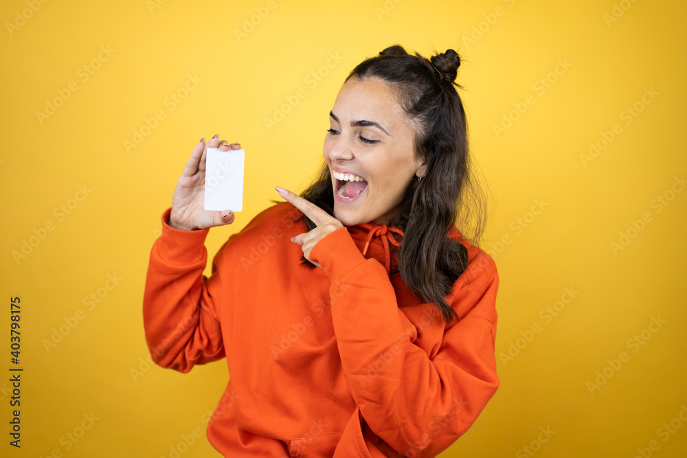 Young beautiful woman wearing sweatshirt over isolated yellow background smiling and holding white card Young beautiful woman wearing sweatshirt over isolated yellow background smiling and holding white card