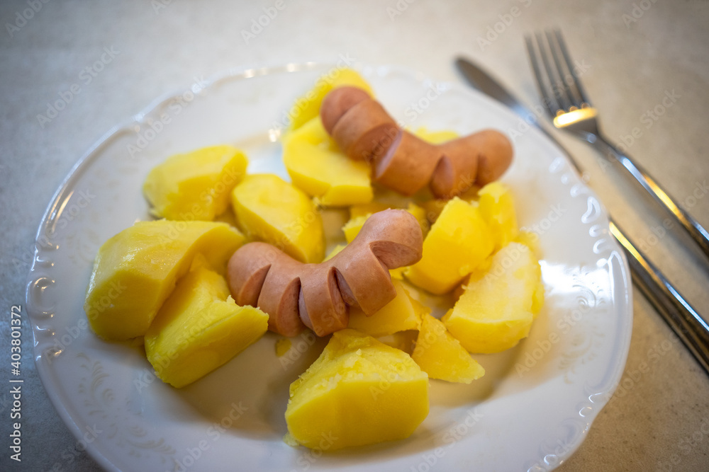 Sausages and potatoes on a white plate on the table, next to a knife and fork Sausages and potatoes on a white plate on the table, next to a knife and fork