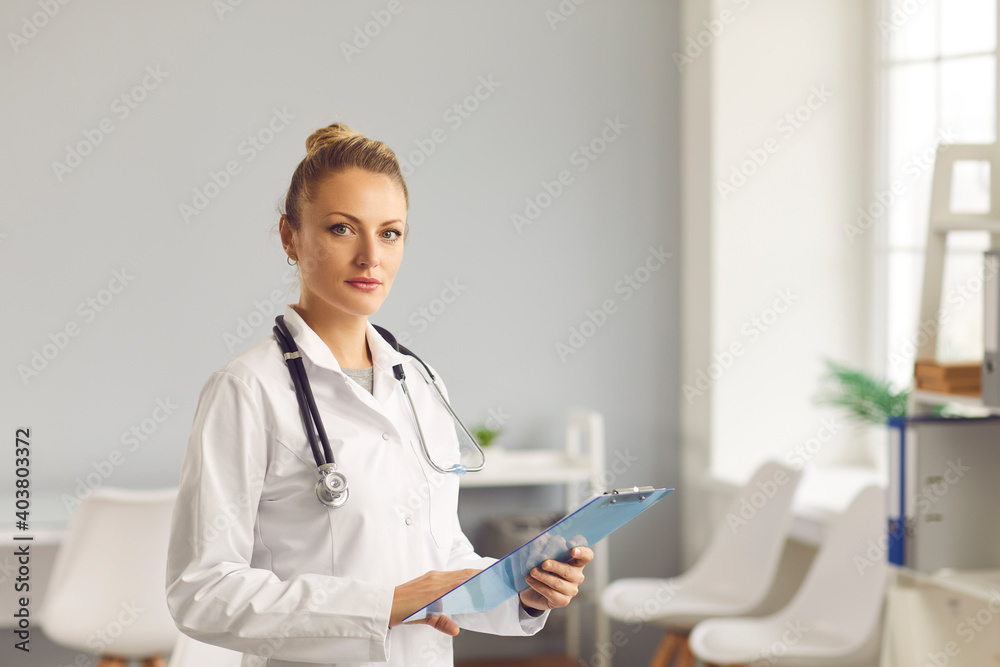 Portrait of a confident young female doctor standing with a clipboard in her hands in the office of a modern medical center. Concept of articles about health care. Banner. Website. Portrait of a confident young female doctor standing with a clipboard in her hands in the office of a modern medical center. Concept of articles about health care. Banner. Website.