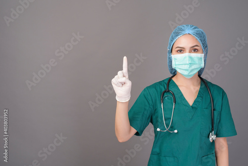 young confident woman doctor in green scrubs is wearing surgical mask over grey background studio