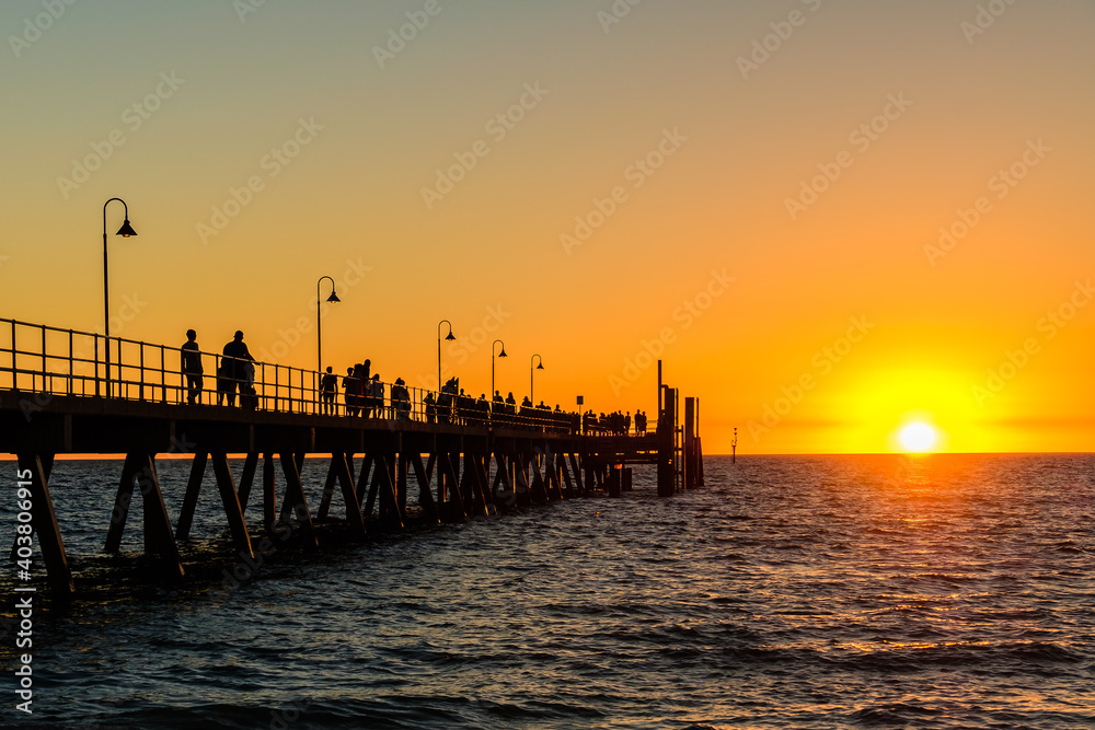 Crowds of people walking along Glenelg Beach jetty at sunset time during a summer evening