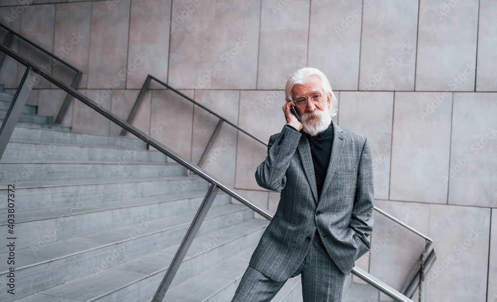 Senior businessman in formal clothes, with grey hair and beard is outdoors talks by phone Senior businessman in formal clothes, with grey hair and beard is outdoors talks by phone
