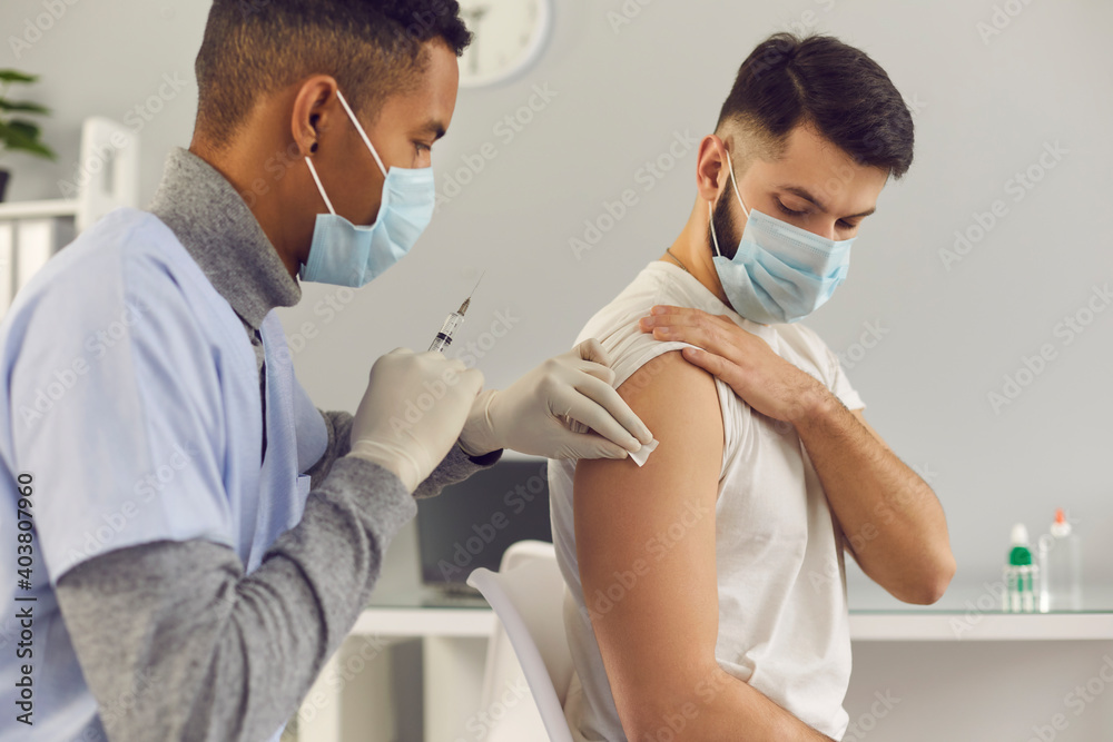 Obraz premium Young man getting flu or Covid-19 antiviral shot during vaccination campaign. African-American doctor in face mask holding syringe and cleaning skin on patient's arm before antivirus vaccine injection