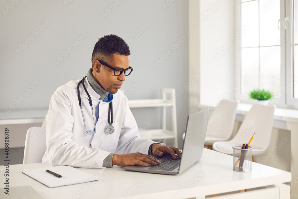 Young African-American doctor sitting at desk, using laptop computer, doing medical research on Internet, filling in electronic health records or giving consultation to patient on online eHealth chat Young African-American doctor sitting at desk, using laptop computer, doing medical research on Internet, filling in electronic health records or giving consultation to patient on online eHealth chat