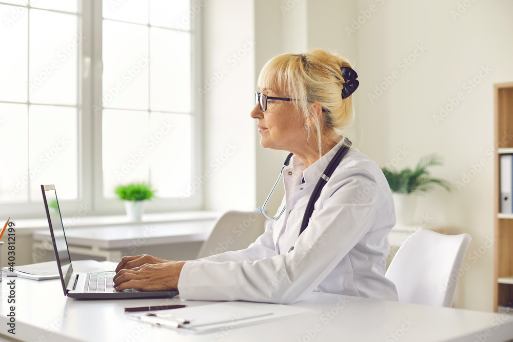 Side view of a female doctor sitting at her workplace in a hospital office typing on a laptop. Older woman in a medical uniform works in a modern medical clinic. Concept of employment at work.