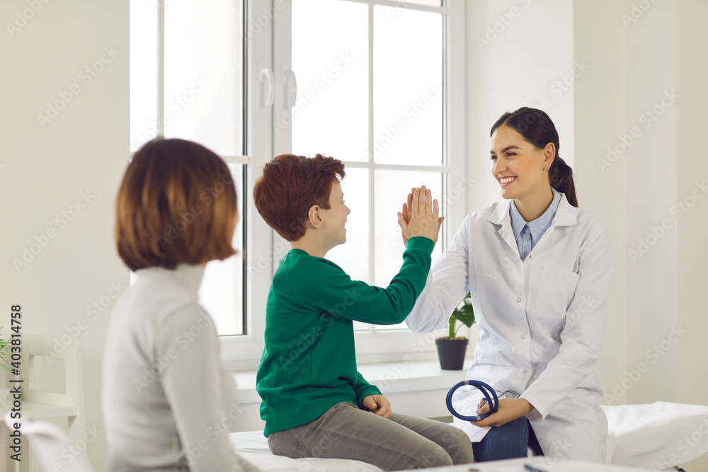 Obraz premium Happy boy patient giving five to smiling woman pediatrician in medical uniform with sitting mother nearby during visit in medical clinic office. Medicare for kids and healthcare concept