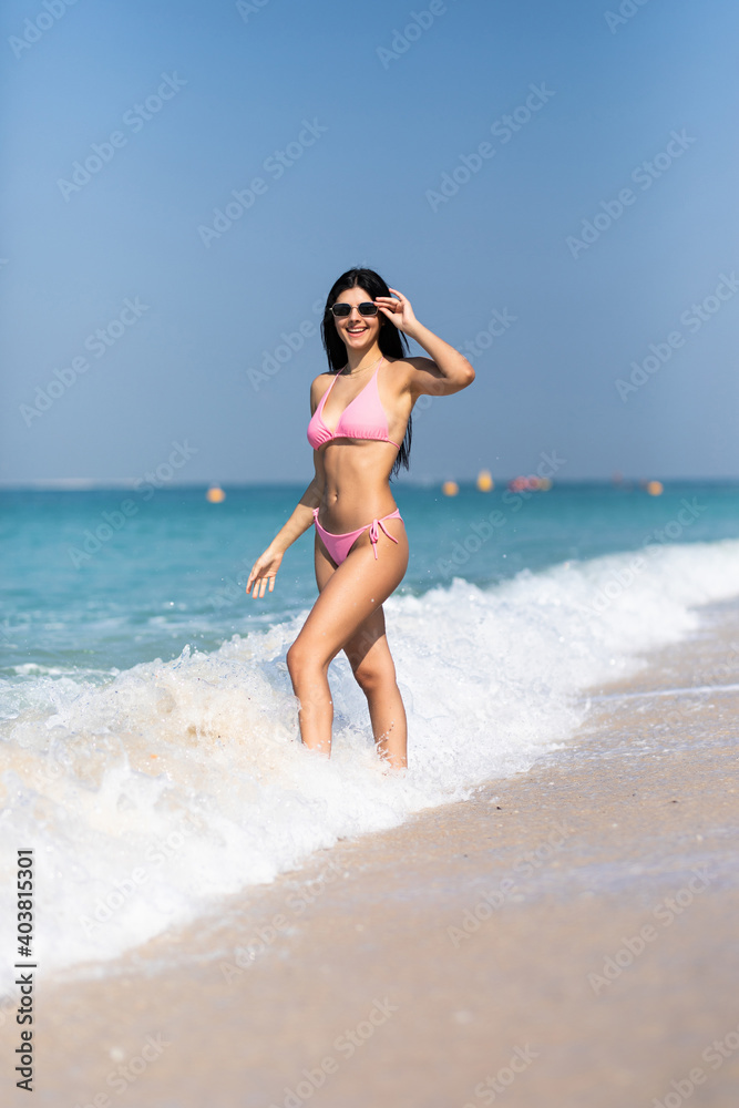 Portrait of young woman in pink bikini on tropical beach looking at camera. Beautiful girl in swimwear with copy space. Summer vacation and tanning concept.