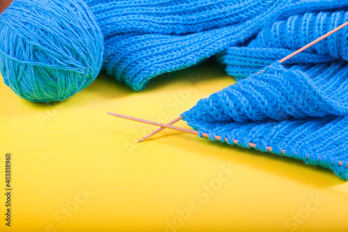 Knitting of a blue scarf with knitting needles and a clew lie on a yellow background.