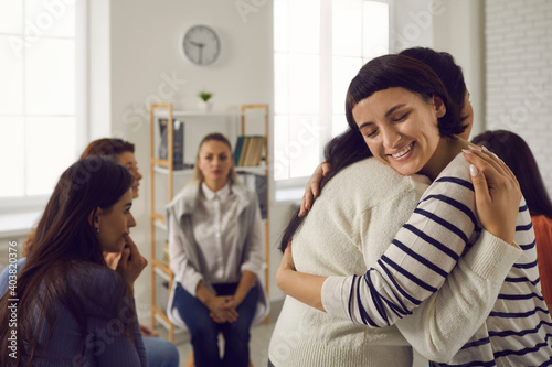 Two happy candid young women hugging each other in support group meeting or therapy session. Concepts of psychological help, overcoming problems together, addiction treatment, empathy and forgiveness