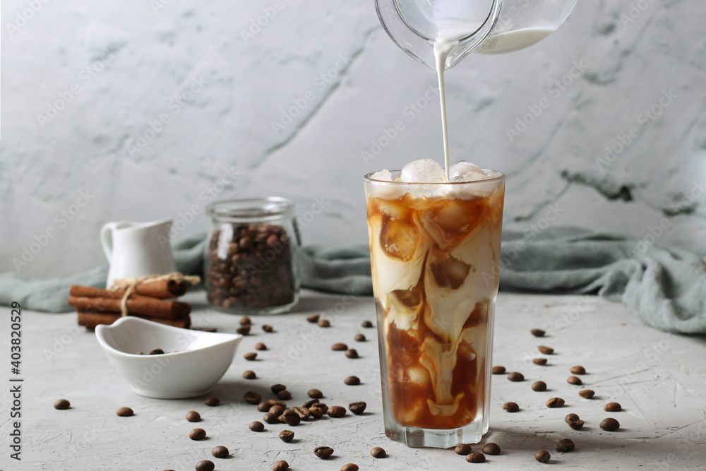 Cold coffee in a tall glass with cream being poured into it showing the texture of the drink. Light gray background, Horizontal format Cold coffee in a tall glass with cream being poured into it showing the texture of the drink. Light gray background, Horizontal format