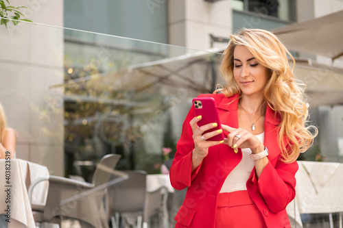 Beautiful blonde woman in red classic smart-casual outfit outdoors near hi-tech business building. She working on her phone. Space for text.