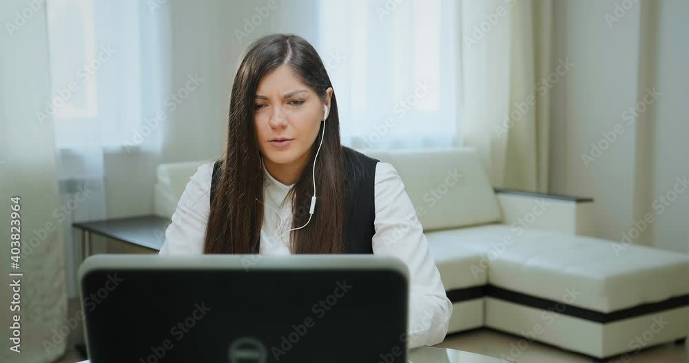 Active brunette with long loose flowing hair in white headphones talks to friends into microphone looking into laptop at home Active brunette with long loose flowing hair in white headphones talks to friends into microphone looking into laptop at home