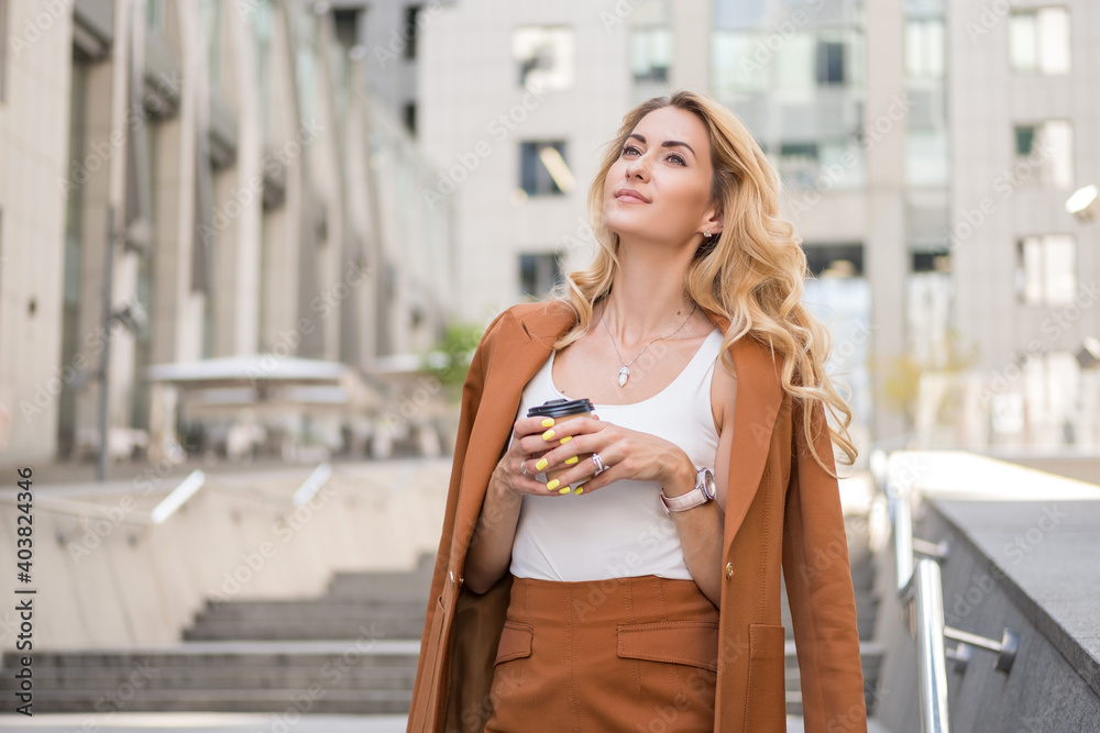 Beautiful blonde woman in brown classic smart-casual outfit outdoors near hi-tech business building. She drinks coffee in to go cup and enjoy summer warmth. Space for text.