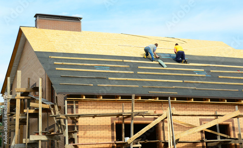 Photos A close-up of a brick house under construction with the roofing contractors on unfinished rooftop installing roofing waterproofing rubberized asphalt underlayment before asphalt shingles installation