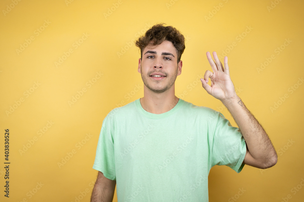 Handsome man wearing a green casual t-shirt over yellow background doing ok sign with fingers and smiling, excellent symbol Handsome man wearing a green casual t-shirt over yellow background doing ok sign with fingers and smiling, excellent symbol