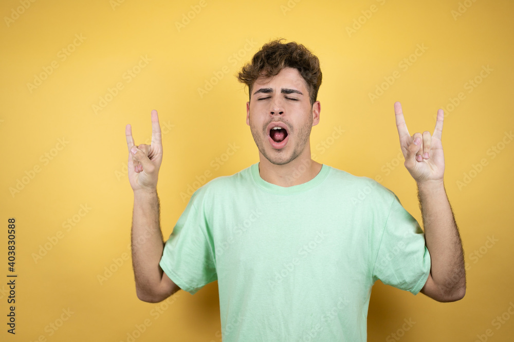Handsome man wearing a green casual t-shirt over yellow background shouting with crazy expression doing rock symbol with hands up Handsome man wearing a green casual t-shirt over yellow background shouting with crazy expression doing rock symbol with hands up