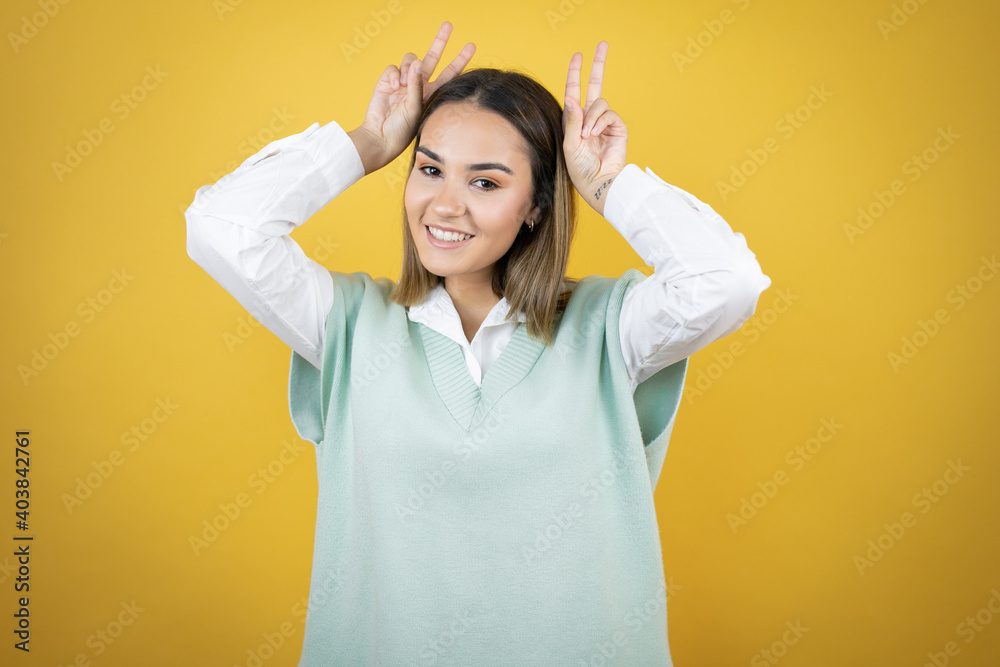 Pretty young woman standing over yellow background Posing funny and crazy with fingers on head as bunny ears, smiling cheerful Pretty young woman standing over yellow background Posing funny and crazy with fingers on head as bunny ears, smiling cheerful