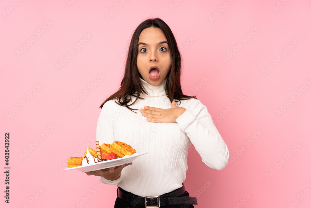 Young brunette woman holding waffles over isolated pink background surprised and shocked while looking right