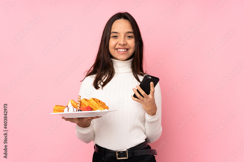 Young brunette woman holding waffles over isolated pink background holding coffee to take away and a mobile Young brunette woman holding waffles over isolated pink background holding coffee to take away and a mobile