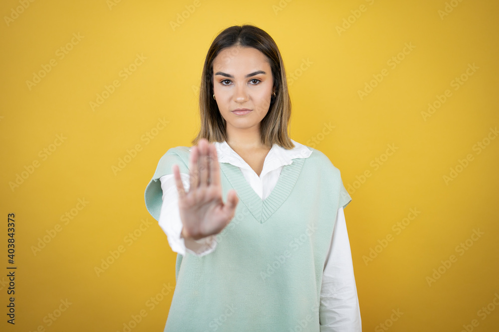 Pretty young woman standing over yellow background serious and doing stop sing with palm of the hand. Pretty young woman standing over yellow background serious and doing stop sing with palm of the hand.
