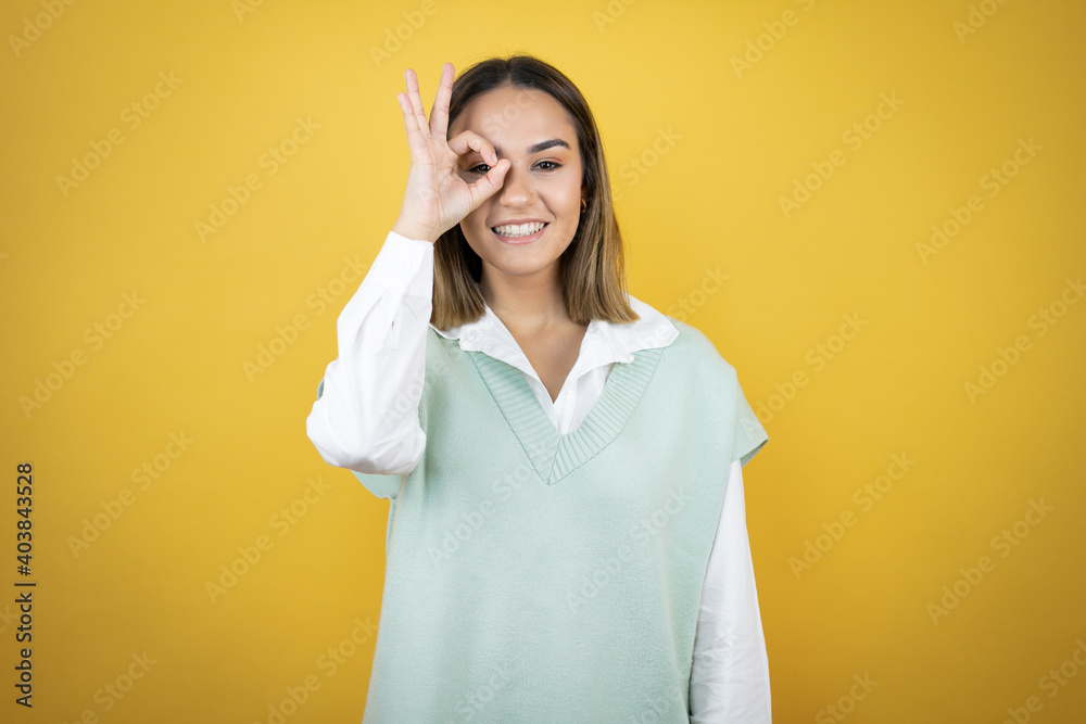 Pretty young woman standing over yellow background doing ok gesture shocked with smiling face, eye looking through fingers Pretty young woman standing over yellow background doing ok gesture shocked with smiling face, eye looking through fingers