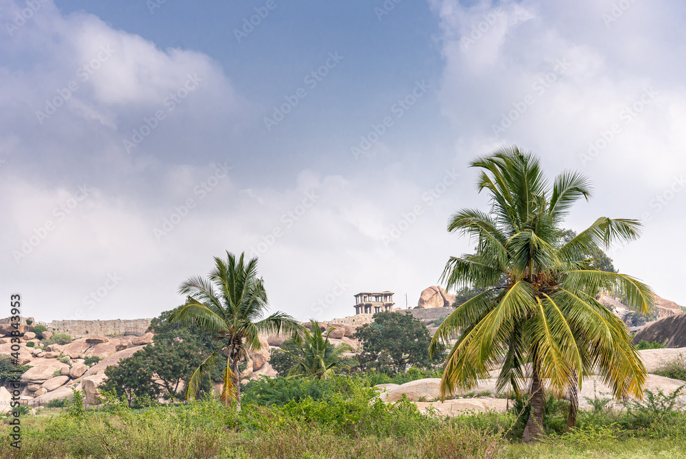 Hampi, Karnataka, India - November 5, 2013: Lakshmi Narasimha Temple. Landscape with ruin on hill and brown rock boulders under blue sky with green foliage and grass up front.
