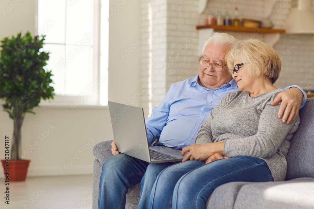 Loving happy elderly couple sitting together on sofa, hugging and watching movie online on laptop at home. Elderly people happy lifestyle and modern technologies concept