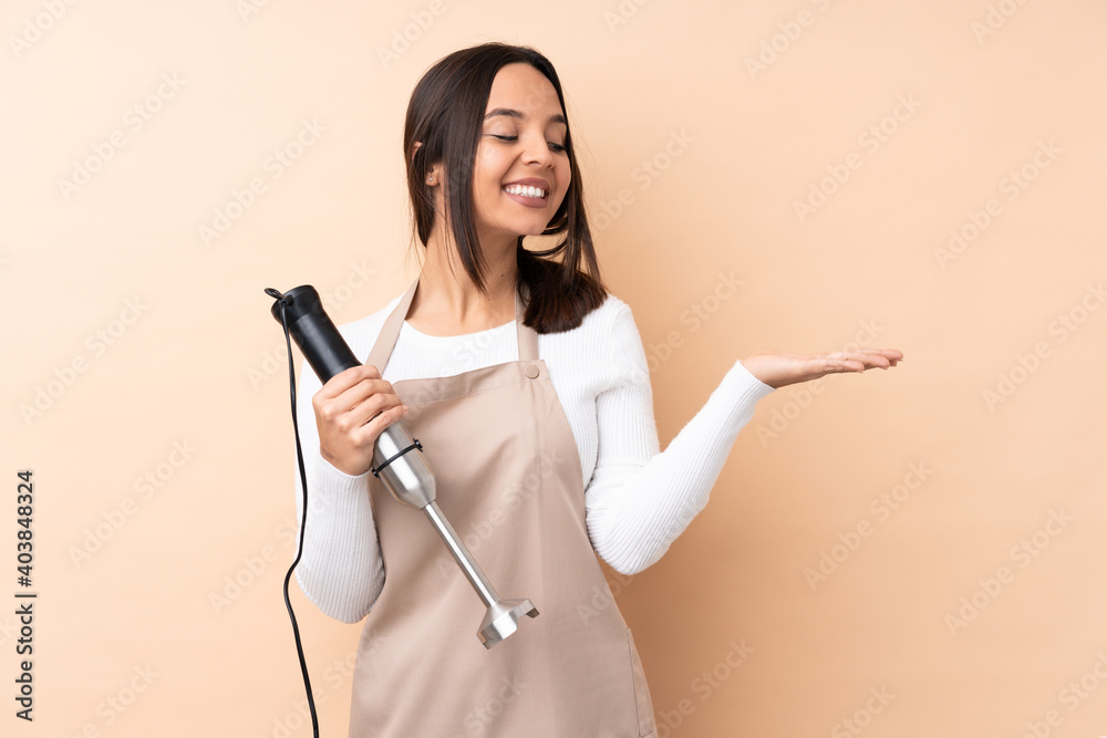 Young brunette girl using hand blender over isolated background holding copyspace imaginary on the palm to insert an ad Young brunette girl using hand blender over isolated background holding copyspace imaginary on the palm to insert an ad