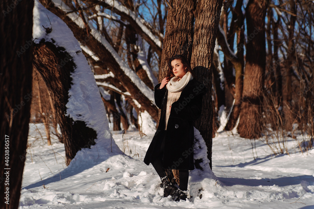 Serious girl in the coat and white scarf leaning on the tree in the winter forest. Sunny winter day