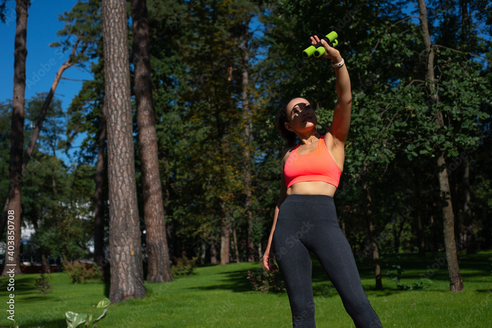 Young sporty woman raising her arm with dumbbells up ad looking at it standing in a city park