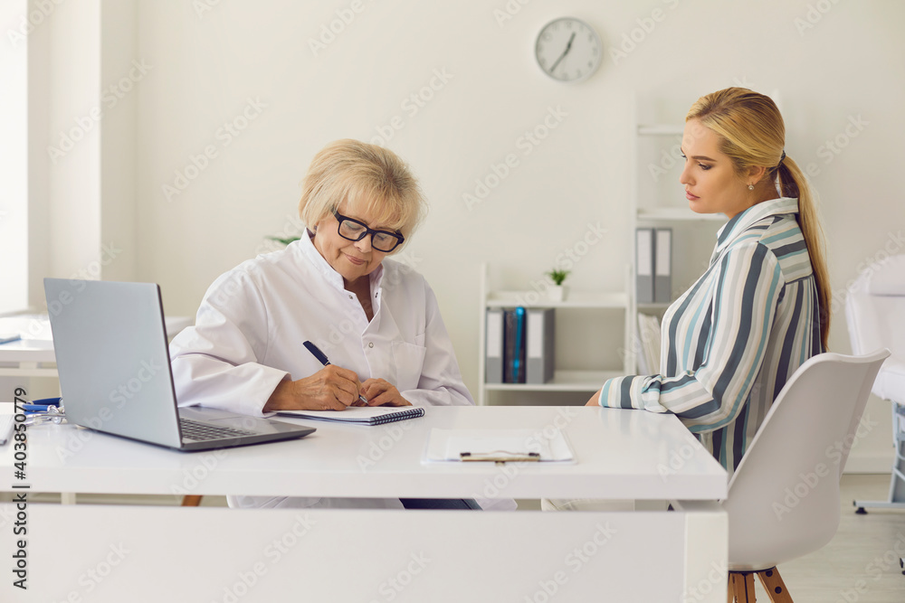 Mature doctor writing prescription for female patient sitting at table in modern hospital office. Senior physician or gynecologist prescribing treatment for young woman Mature doctor writing prescription for female patient sitting at table in modern hospital office. Senior physician or gynecologist prescribing treatment for young woman